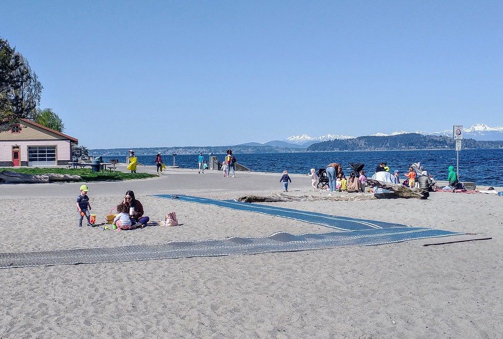 A bustling Alki Beach scene with beachgoers, playful children, and the majestic Olympic Mountains in the distance.